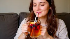 Žena pije ovocný čaj s brčkem, šťastně sedí na gauči v obývacím pokoji.