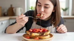Žena jí toast s čokoládou a jahodami u stolu v kuchyni, domácí sladká snídaně.