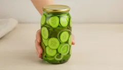 Zavařovací sklenice s nakládanými okurkami na světlém stole, ruce držící sklenici.